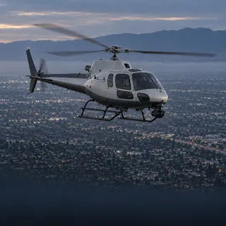 Illustration of an unmarked grey news helicopter over the Los Angeles street grid at dusk. No specific aircraft is depicted.