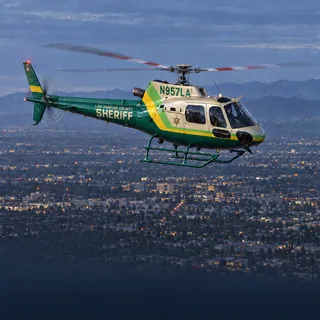 Illustration of a Los Angeles County Sheriff's Department patrol Airbus AS350 in green and cream, registration N957LA, over Los Angeles at dusk.