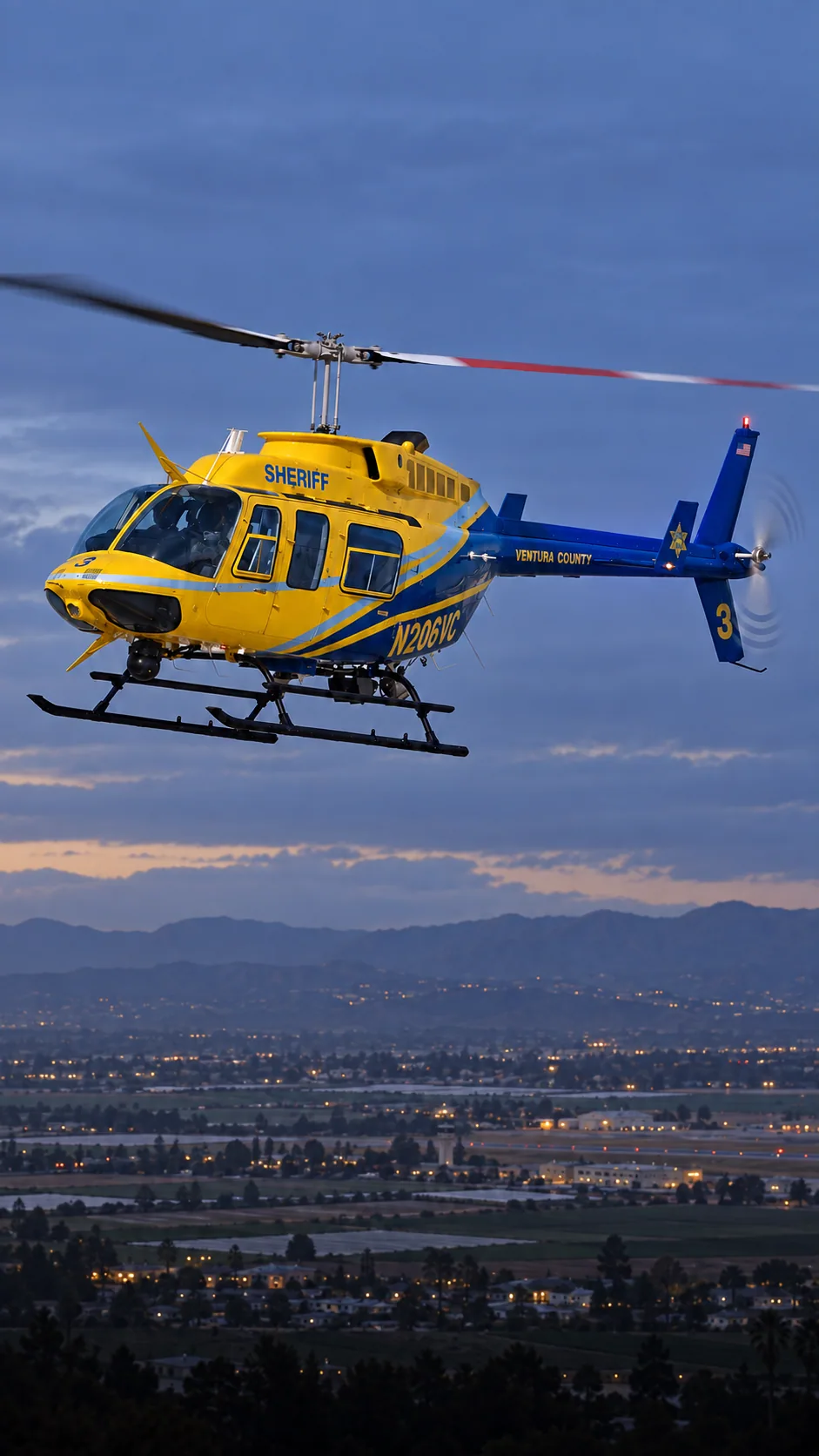 Illustration of N206VC, a Bell 206L-1 LongRanger of the Ventura County Sheriff — nose LEFT with port side visible enough to read low cabin registration — over Camarillo, Ventura County.