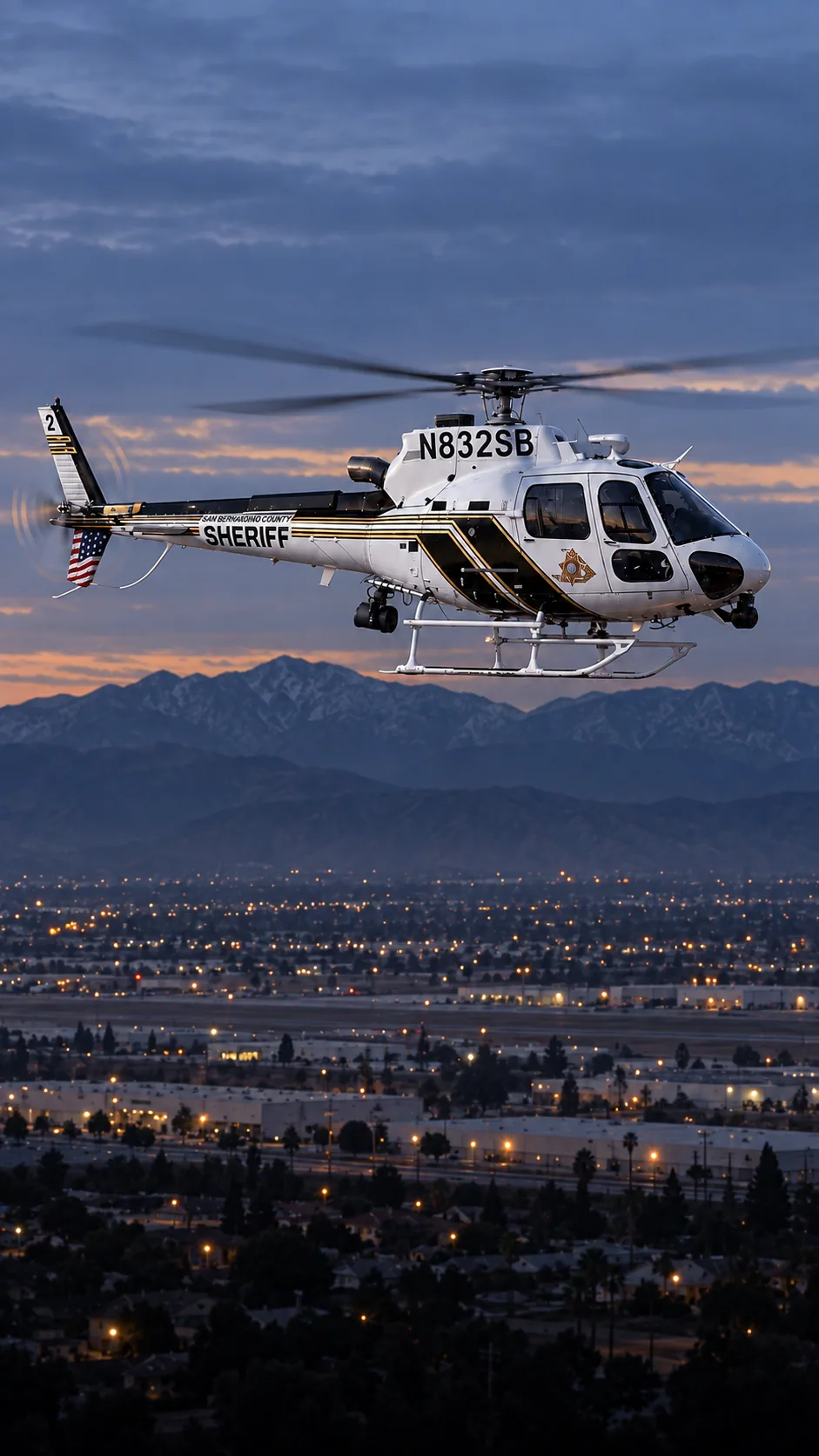 Illustration of N832SB, an Airbus AS350 of the San Bernardino County Sheriff — white cabin, black diagonal side panels and top tail boom, thin gold and black stripes — over San Bernardino and Highland at the foot of the San Bernardino Mountains.