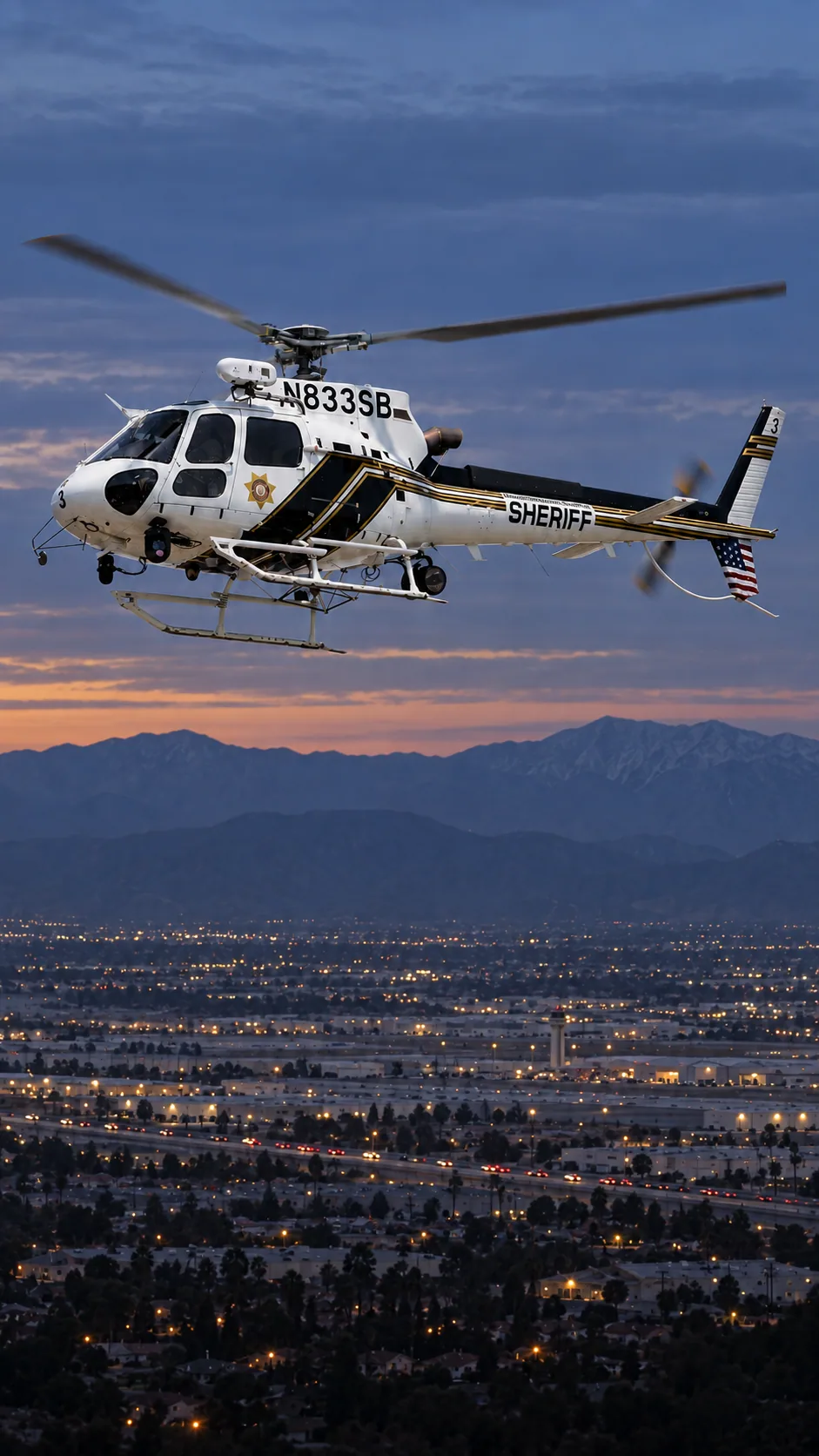 Illustration of N833SB, an Airbus AS350 of the San Bernardino County Sheriff — white cabin, black diagonal side panels and top tail boom, thin gold and black stripes — over San Bernardino and Highland at the foot of the San Bernardino Mountains.