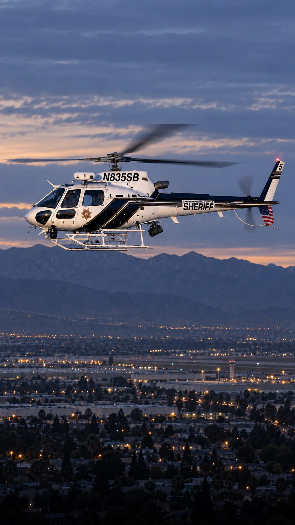 Illustration of N835SB, an Airbus AS350 of the San Bernardino County Sheriff — white cabin, black diagonal side panels and top tail boom, thin gold and black stripes — over San Bernardino and Highland at the foot of the San Bernardino Mountains.