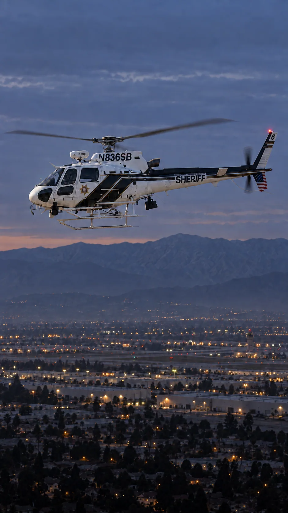Illustration of N836SB, an Airbus AS350 of the San Bernardino County Sheriff — white cabin, black diagonal side panels and top tail boom, thin gold and black stripes — over San Bernardino and Highland at the foot of the San Bernardino Mountains.