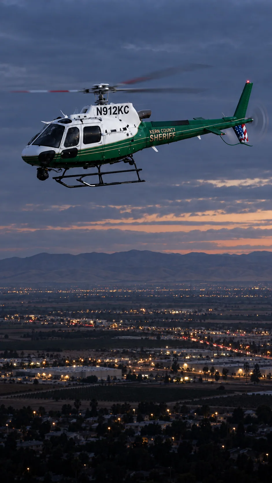Illustration of N912KC, an Airbus AS350 of the Kern County Sheriff — white cabin and cowling, green lower fuselage and tail, thin gold pinstripes — over Bakersfield, Kern County, seen from the Meadows Field side of town.
