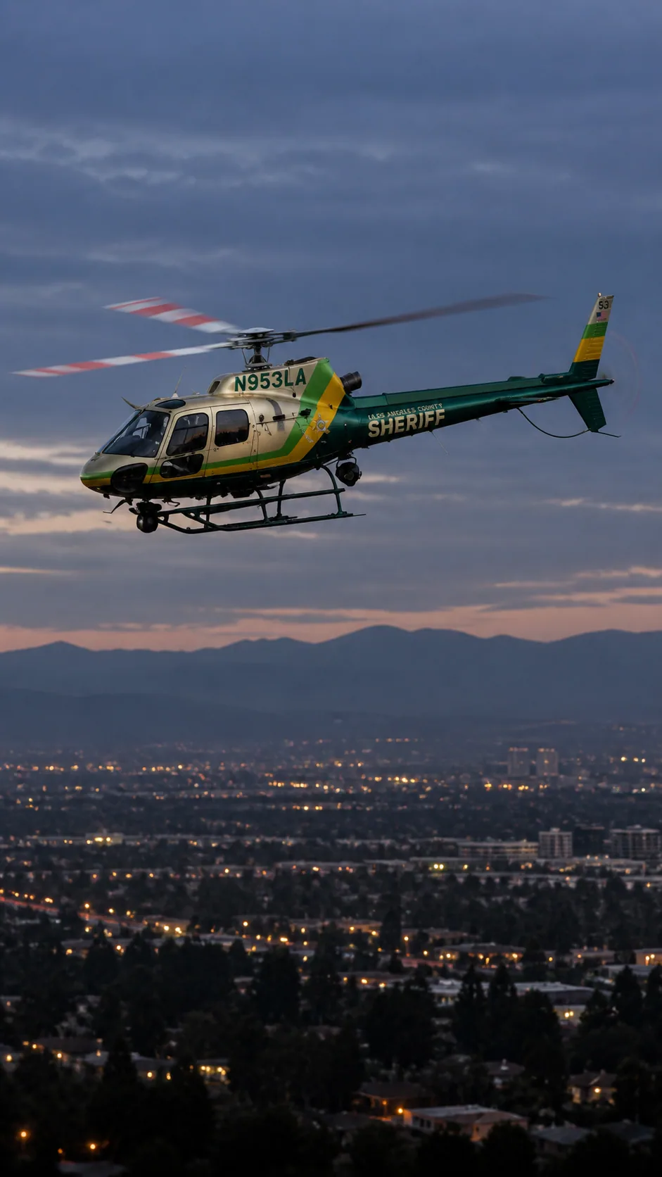 Illustration of N953LA, an Airbus AS350 B2 of Sheriff Air Support — metallic champagne/gold cabin, emerald green tail and belly, lime green and yellow diagonal bands — over the Los Angeles basin at dusk.