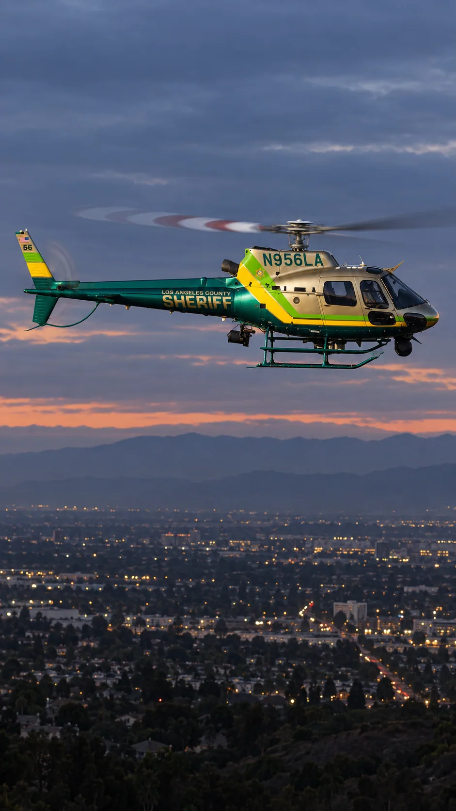 Illustration of N956LA, an Airbus AS350 B2 of Sheriff Air Support — metallic champagne/gold cabin, emerald green tail and belly, lime green and yellow diagonal bands — over the Los Angeles basin at dusk.