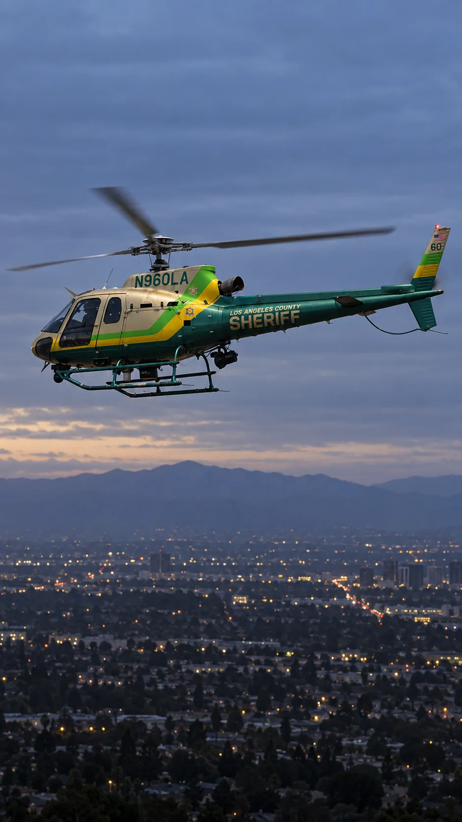 Illustration of N960LA, an Airbus AS350 B2 of Sheriff Air Support — metallic champagne/gold cabin, emerald green tail and belly, lime green and yellow diagonal bands — over the Los Angeles basin at dusk.