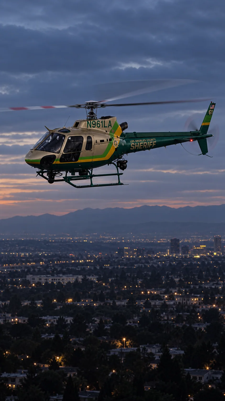 Illustration of N961LA, an Airbus AS350 B2 of Sheriff Air Support — metallic champagne/gold cabin, emerald green tail and belly, lime green and yellow diagonal bands — over the Los Angeles basin at dusk.