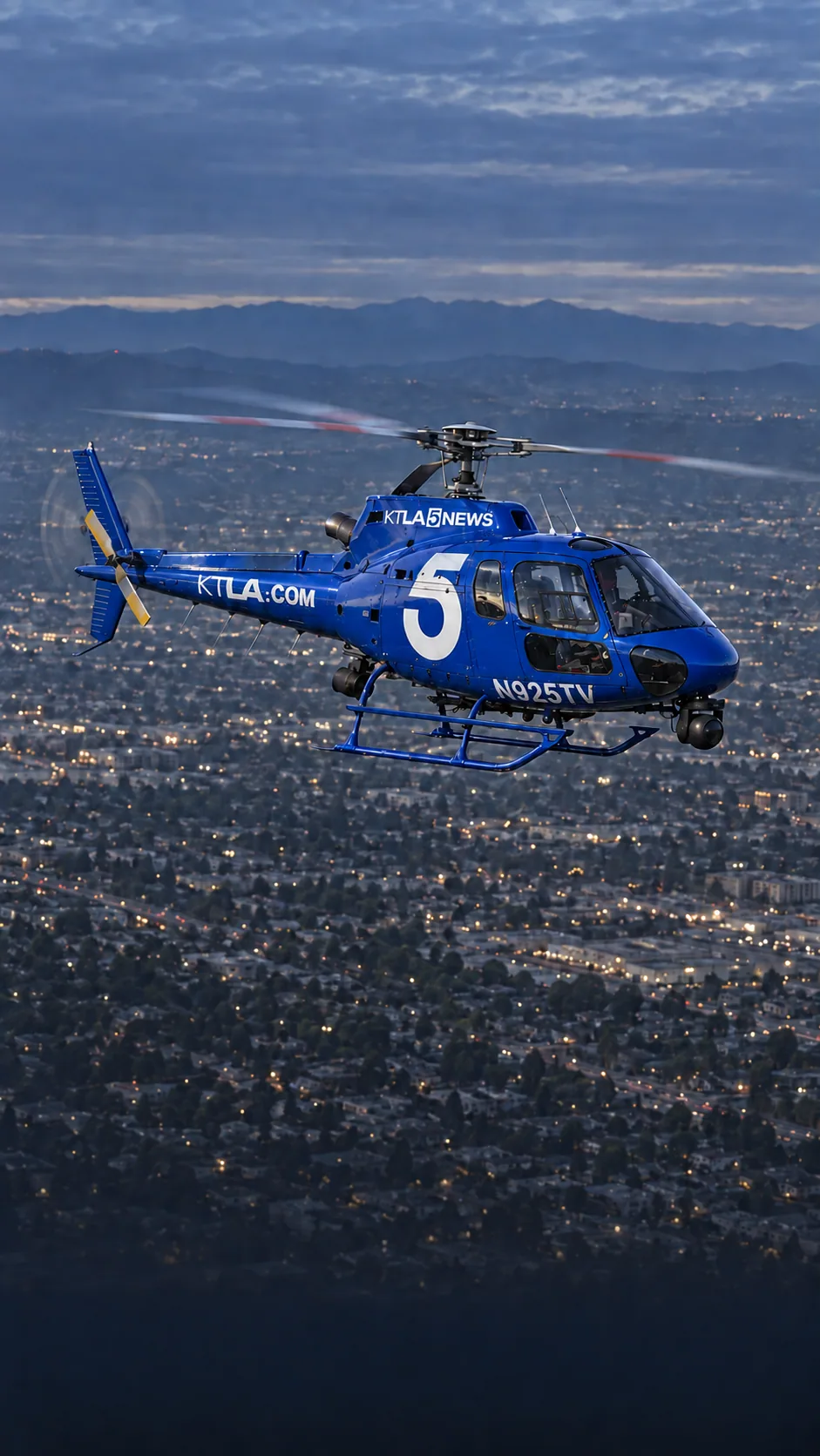 Illustration of Sky5, KTLA's royal-blue Airbus AS350 news helicopter with a large white 5, registration N925TV, over Los Angeles at dusk.